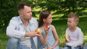 Caucasian family eats apples on picnic blanket under trees. Parents and two children are nibbling on fruit in open air. Dog lies casually near basket, nibbling on apple. Family Day, family leisure - Powered by Shutterstock - Get 15% off with code: PIKWIZARD15