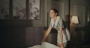 Smiling Young Hotel Housekeeper In Neat Uniform Carefully Makes Bed, Spreading Fresh, Crisp White Linens. Soft Daylight Fills Room. Concept of Cleanliness, Comfort High Standard Hospitality Service. - Powered by Shutterstock - Get 15% off with code: PIKWIZARD15
