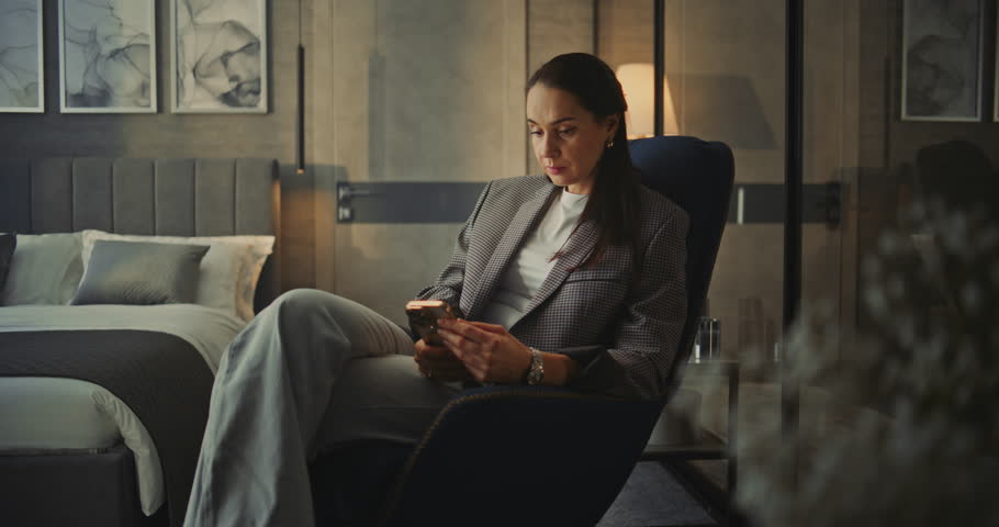 Stylish Woman With Long Dark Hair Sits in Soft Armchair in Hotel Room With Phone in Hands. Woman Checks Work Email and Responds to Messages. Concept Remote Work, Business Routine, Connection in World.