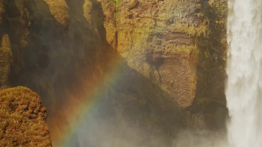 wide angle shot of the famous Skógafoss waterfall in Iceland. The powerful cascade of water plunges over a massive cliff, creating a misty spray at the base. A vibrant rainbow arches across the mist,