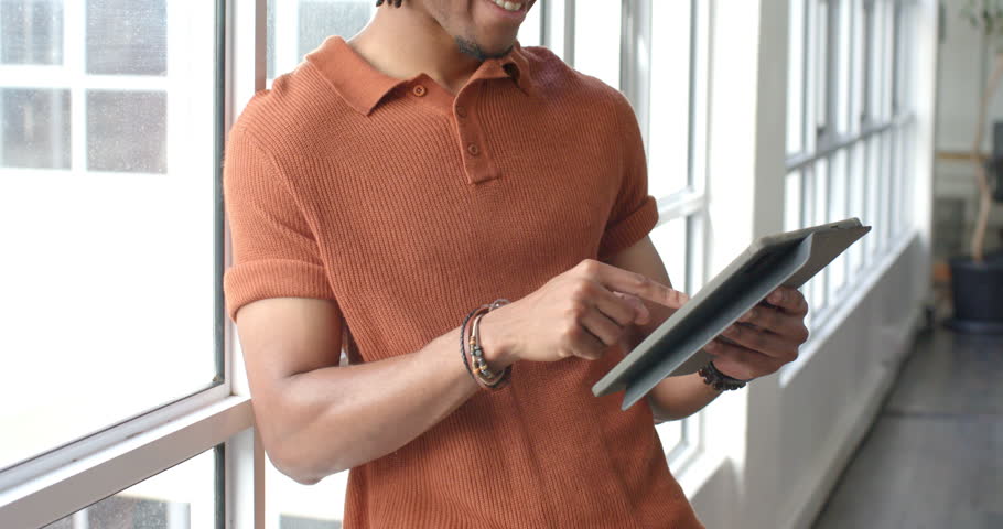 African American man scrolling tablet while leaning on windowsill in office reviewing content. Technology, future, innovation, modern, productivity, workspace, casual
