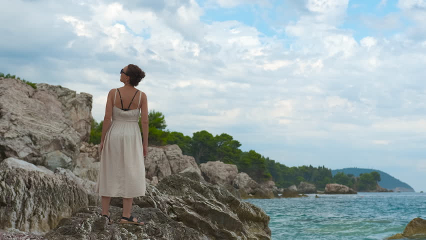 Woman in a dress standing on a rocky shore looking at the sea. Young woman in a summer dress and sunglasses standing on a rocky coast and admiring the beautiful seascape on a cloudy day