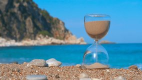 Hourglass sand measuring time on a sunny beach. Close-up of an hourglass on a sandy beach with pouring sand measuring the passing time against a beautiful blue sea and rocky cliff background - Powered by Shutterstock - Get 15% off with code: PIKWIZARD15