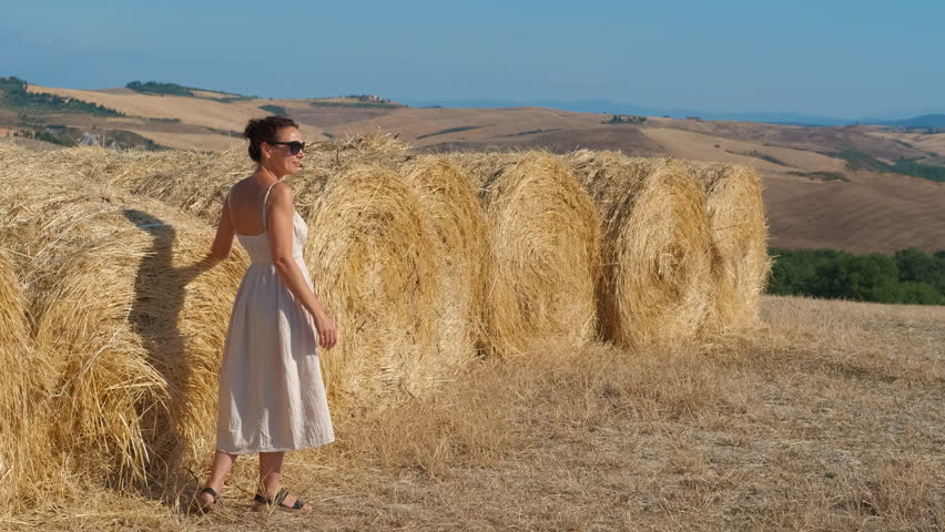 Woman touching hay bales in a summer wheat field. Beautiful woman in a dress walking in a harvested wheat field, touching a large round hay bale while enjoying the scenic tuscan countryside landscape