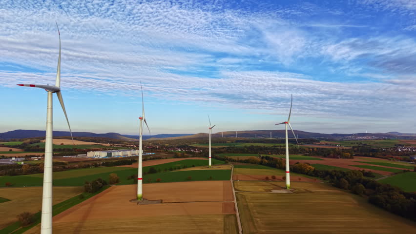 Majestic wind turbines rise above green fields and golden crops, creating a powerful contrast against the blue sky and fluffy white clouds in the serene landscape.