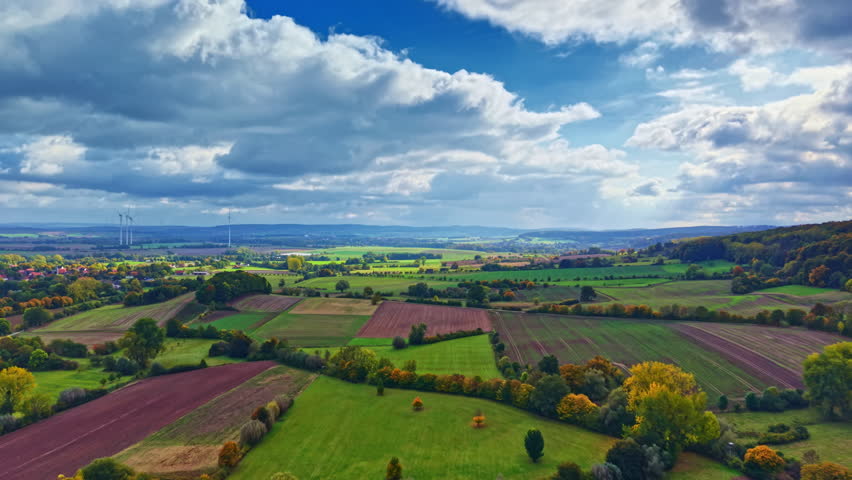 A breathtaking view of diverse farmland showcases green fields, purple crops, and scattered trees under a dynamic sky filled with fluffy clouds, all reflecting the beauty of autumn.