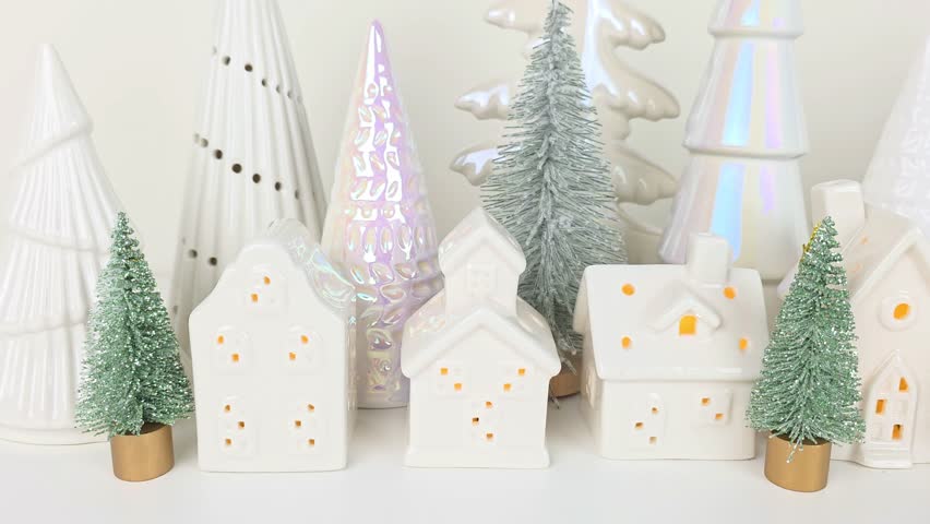 Tiny Christmas village with lit white houses and tree decor in the background. Slow panning motion across a white shelf.