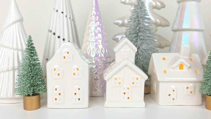 Tiny Christmas village with lit white houses and tree decor in the background. Slow close up panning motion across a white shelf.