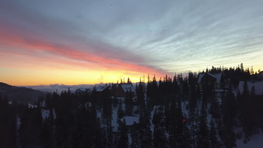 Sunset over snowy mountain landscape with silhouetted trees and cabins
