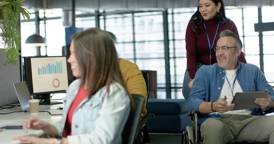 Noticing chat, diverse coworkers gathering around wheelchair user at desk reviewing tablet charts. Collaboration, diversity, teamwork, inclusion, modernoffice, digitaltools, professional