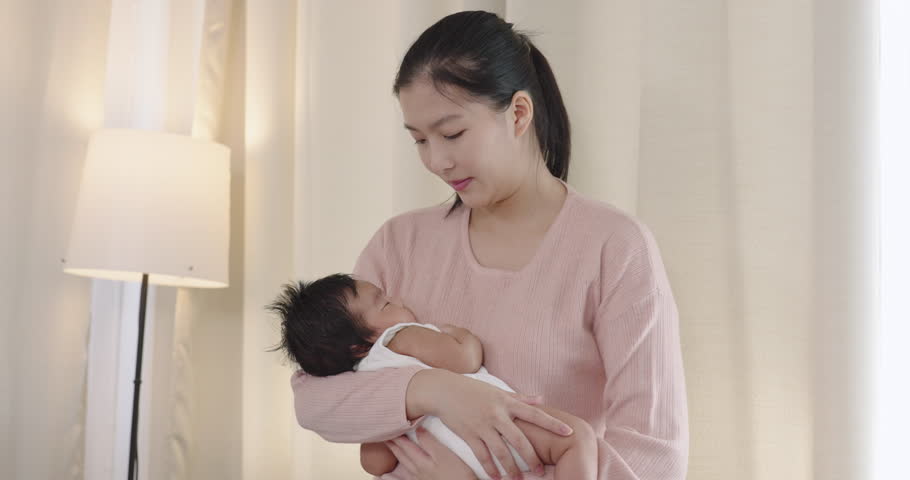 Asian mother looking down at newborn baby boy in arms while standing in softly lit room, warm tender moment of early motherhood and emotional bonding in peaceful home setting