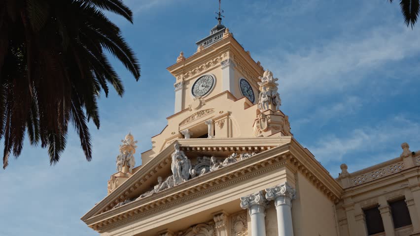 Upward shot of Málaga City Hall, Costa del Sol, Spain, clock tower framed by a tall palm tree against a clear blue sky, capturing elegant Spanish architecture and bright Mediterranean sunlight.