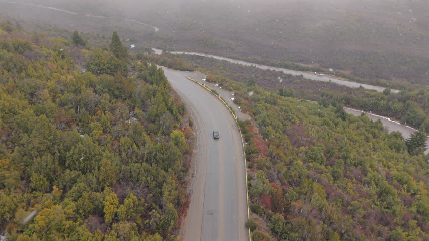 Aerial view shows a lone car following a winding mountain road through Andean forest during a low-visibility snowstorm near Bariloche, with fog and muted autumn colors in northern Patagonia.