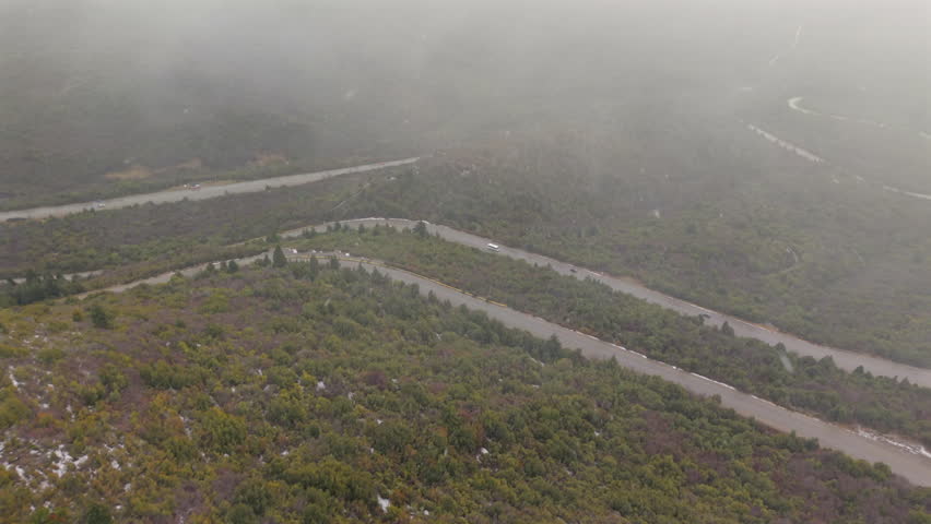 Drone captures winding mountain road carving through Patagonian forest in low visibility as snowstorm sweeps the hills near Bariloche, Argentina—subdued car lights barely visible through swirling mist