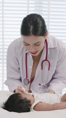 Asian female pediatrician adult examining asian newborn baby lying on bed, gently observing baby's face with warm smile, early checkup in cozy clinic environment, close body interaction