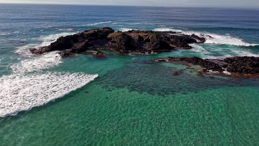 Drone moves backward from rocky islet over turquoise waves toward beach.
