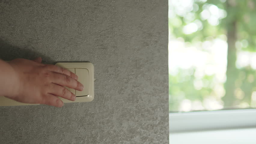 Hand presses switch and roller blinds on windows lower, darkening room. Privacy, security, and comfort in residential buildings.