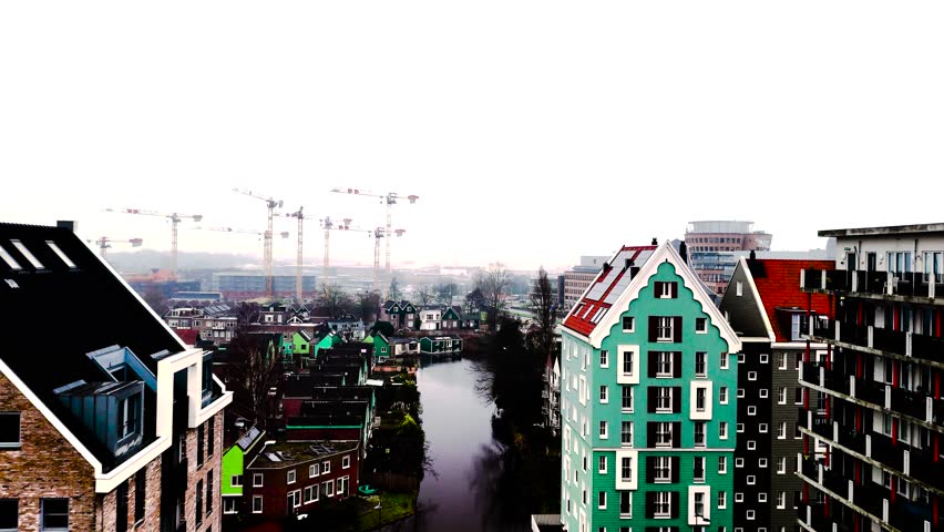 A panoramic view of Amsterdam shows modern buildings with vibrant colors alongside quiet canals. Cranes indicate ongoing construction in the area, blending old and new designs.