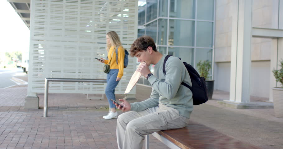 Man holding phone at shelter bench as woman waiting triggers opening sandwich and sipping drink. Urban, casual, lifestyle, modern, technology, communication, leisure