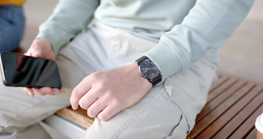 Diverse couple sitting park bench while man glancing at smartphone checking wristwatch to see time. Romantic, casual, lifestyle, modern, leisure, connection, vibrant