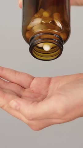 Vertical close-up of a hand full of white capsules while more are being poured from a bottle. Concept of supplements, vitamins and daily healthcare routine.