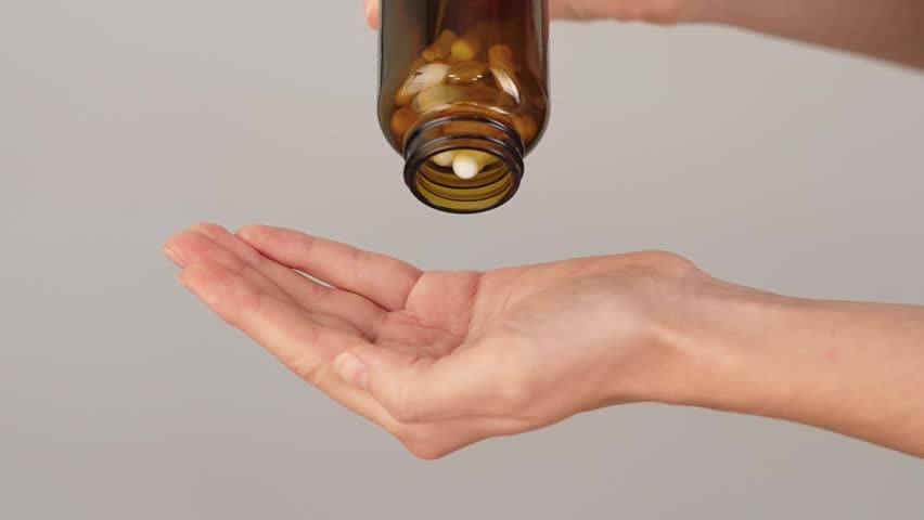 Close-up of a hand full of white capsules while more are being poured from a bottle on a neutral background. Concept of supplements, collagen, probiotics, pain relief medication, pharmaceutical pills.