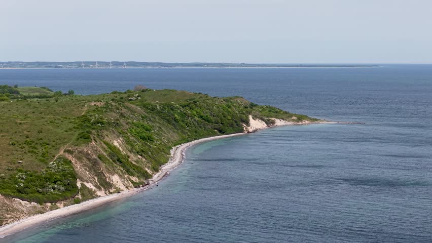 Aerial drone footage of the coastal cliffs near Sletterhage, Denmark, showing steep green hills, rocky shoreline, and calm blue water under bright daylight