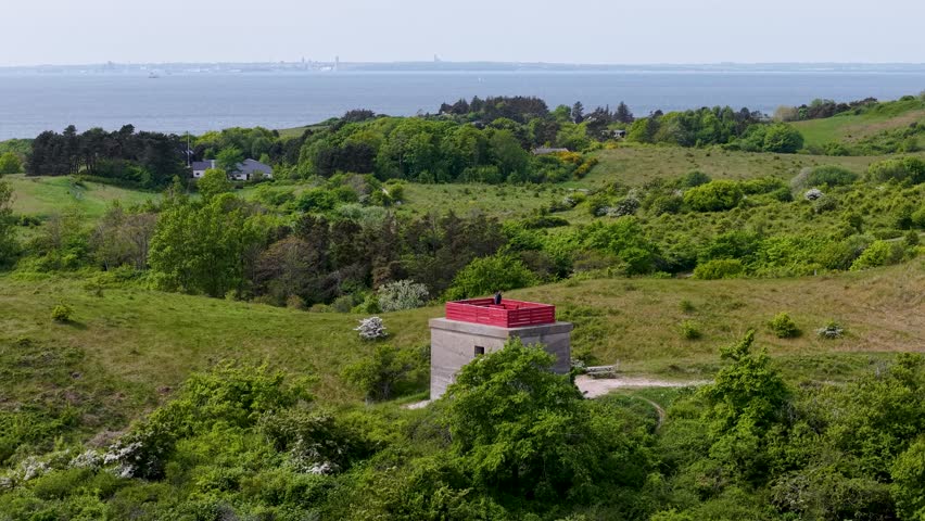 Aerial drone footage of Tyskertårnet, the German World War II bunker tower on the Danish coast, surrounded by green hills and overlooking the sea under cloudy daylight