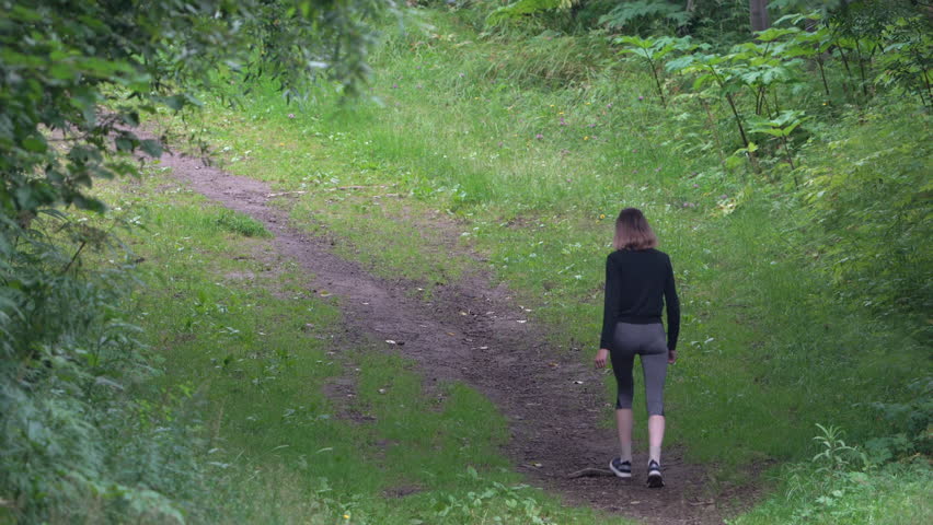 Rear view of fit model during summer sports training walking along pathway in park. Unrecognizable female sportsperson wearing sports clothing - tracksuit jacket and cropped leggings. Zoom in