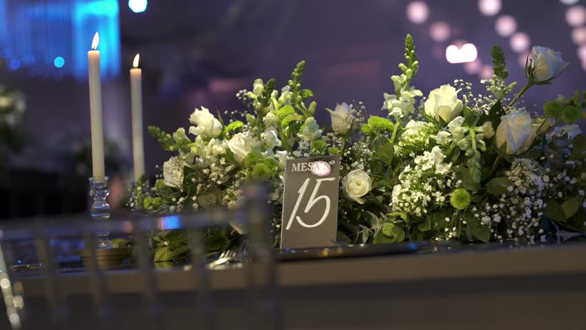 Refined wedding table setup features white roses, baby’s breath, and greenery with two taper candles and a table “Mesa N° 15” card, creating a romantic and festive ambiance under soft string lights.