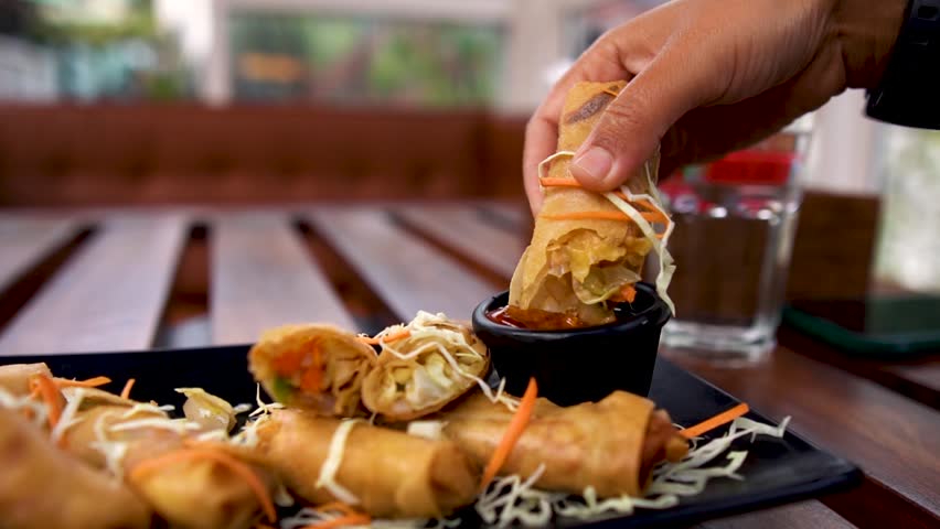 Close up shot of woman hand dip spring roll in sauce at Indian restaurant. Delicious crispy spring rolls filled with savory ingredients. Fried appetizer in plate. Indian woman eating food with hands.