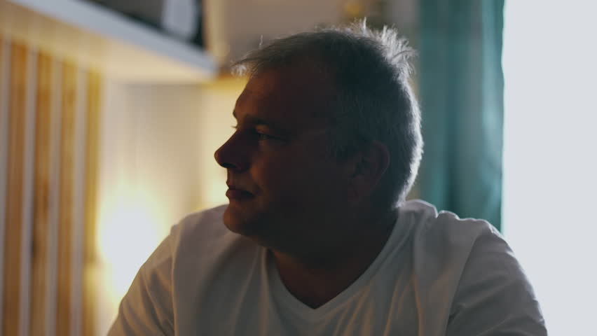 Middle-aged man sitting on bed with hand on forehead, shaking head in disbelief and regret, expressing emotional pain, sorrow, and self-reproach in intimate domestic setting
