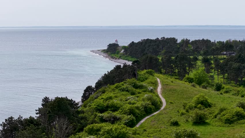 Aerial drone footage of Sletterhage Lighthouse on the coast of Denmark, showing the white lighthouse, rocky shoreline, and surrounding green hills under bright daylight