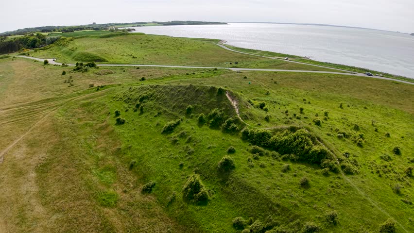 Aerial drone footage of Denmark’s coastal landscape with rolling green hills, grassy terrain, and calm shoreline under cloudy daylight