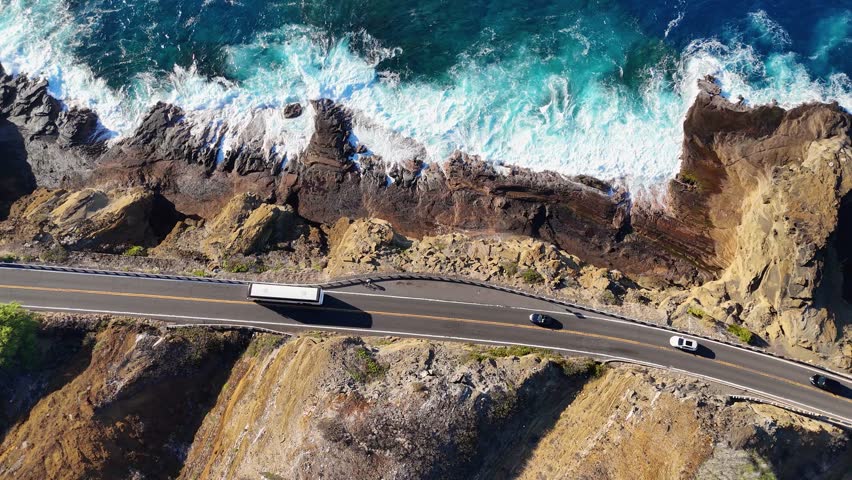 Aerial view of cars traversing a coastal road adjacent to a rocky shoreline with turquoise ocean waves crashing against the cliffs, Honolulu, Hawaii, United States.