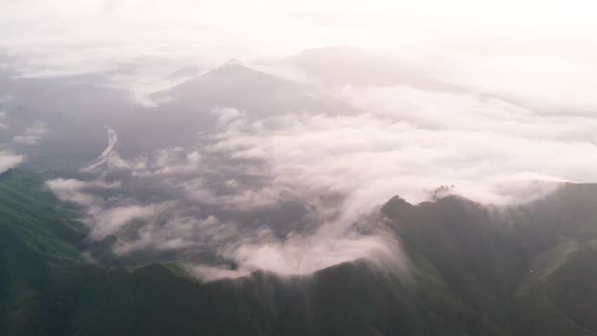 Breathtaking high-altitude aerial view of steep, rugged mountain ridges and peaks. Thick, swirling white clouds and fog roll dramatically through the deep valleys and crests of the mountains below.