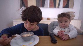 Older brother eating cereal while baby watches curiously from high chair, showing sibling interaction and early social observation during breakfast at home - Powered by Shutterstock - Get 15% off with code: PIKWIZARD15