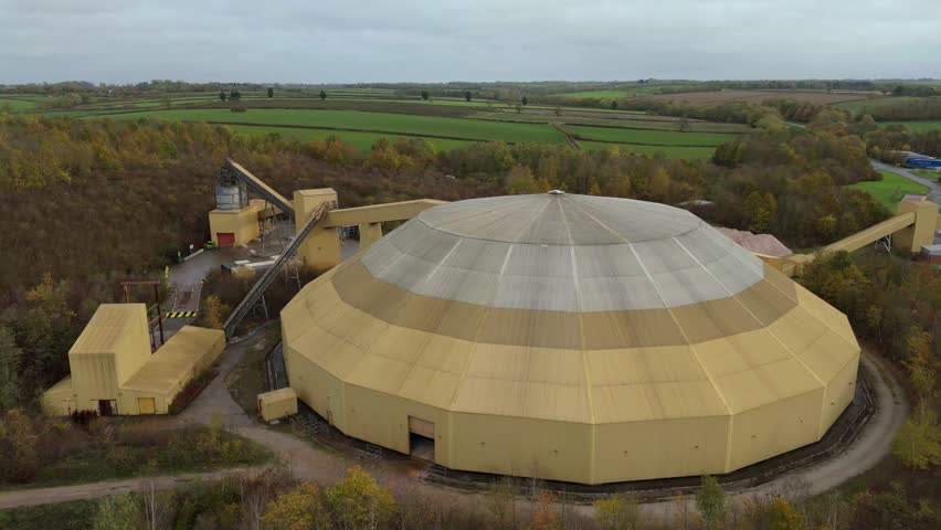 Aerial view of the Sileby Dome, a large, tiered structure with a muted yellow and grey hue, contrasting against the surrounding green fields, Sileby, England, United Kingdom.