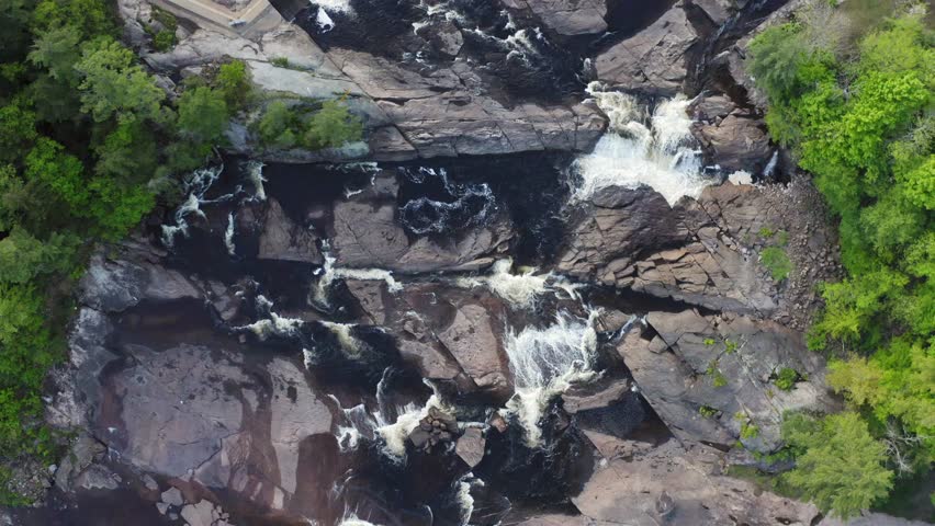 Aerial top view over a wide river flowing down a rocky terrain, surrounded by a green mountain forest