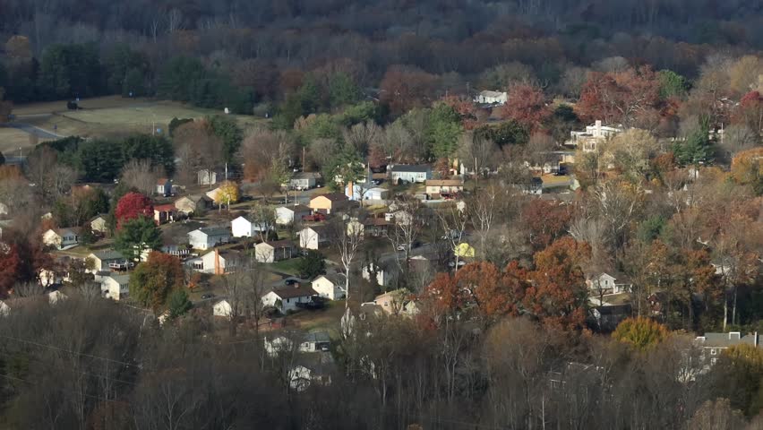 Sweeping aerial panorama of a peaceful American suburban landscape among rolling hills, rows of houses and vibrant autumn trees in midday light, evoking tranquil living and green connectivity.
