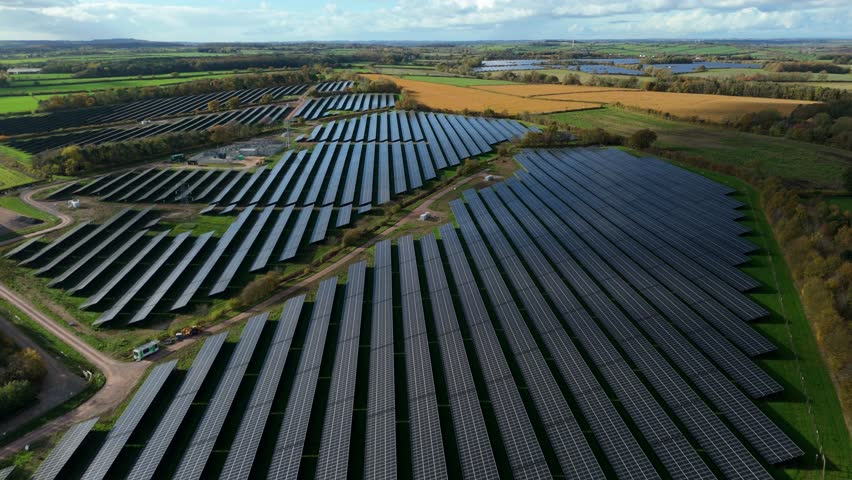 Aerial view of a vast solar panel farm contrasting with green fields, and autumn trees under a blue sky, Southwell, England, United Kingdom.