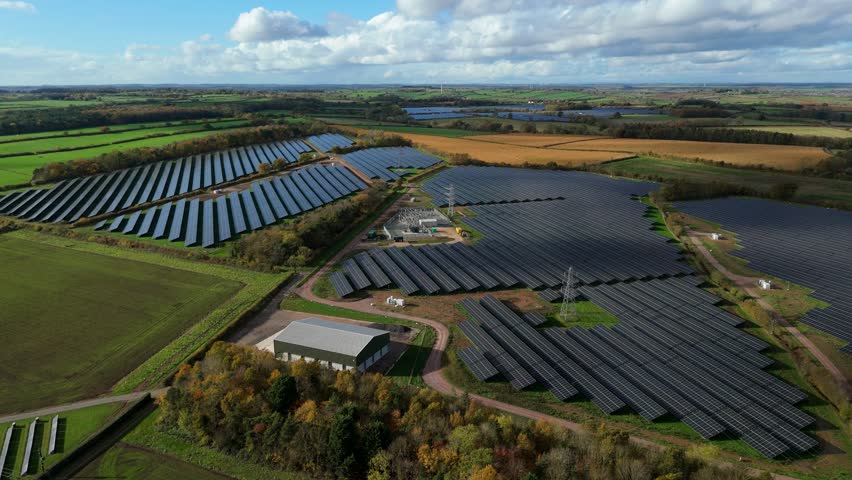 Aerial view of a vast Solar Farm, with rows of panels offset by lush green fields and autumnal trees under a partly cloudy sky, Southwell, England, United Kingdom.