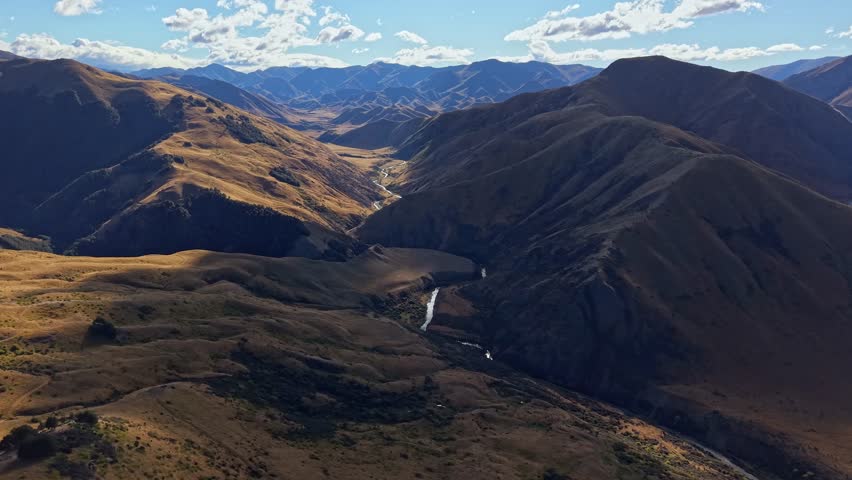 Aerial panning drone shot of Lindis Pass in New Zealand, showing mountain ridges, shadows of clouds and a winding valley under a blue sky.