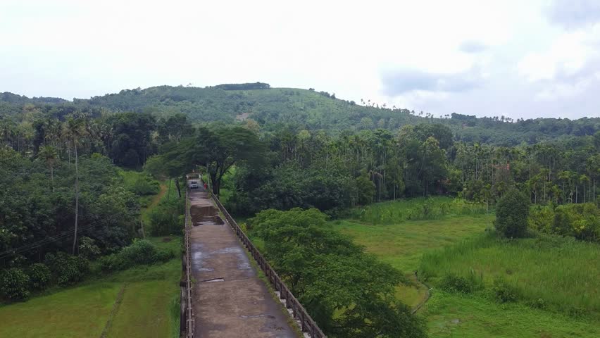 Aerial shot of a white car driving down a narrow, elevated road or bridge crossing a lush green tropical valley, surrounded by dense forest and hills in South India.