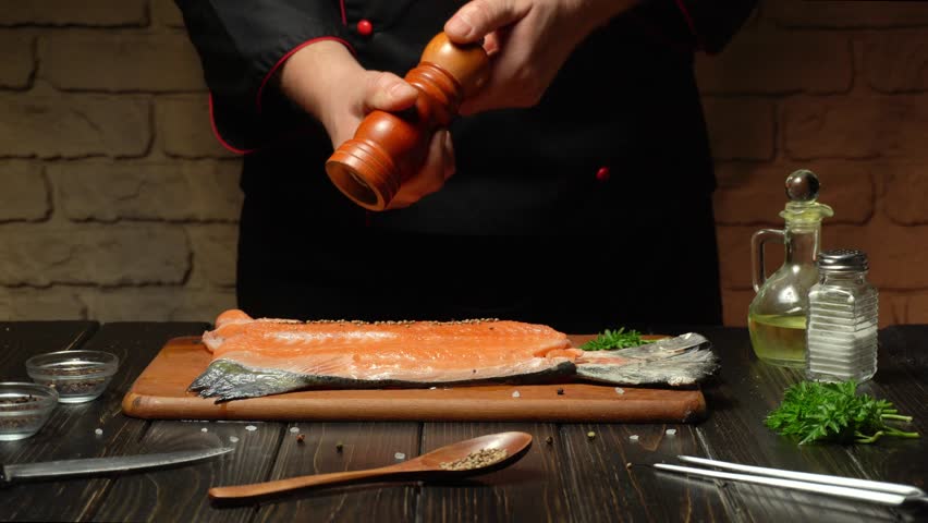 A chef is seasoning a raw salmon fillet with freshly ground pepper in a warm kitchen. Various herbs and seasoning are laid out for enhanced flavor.