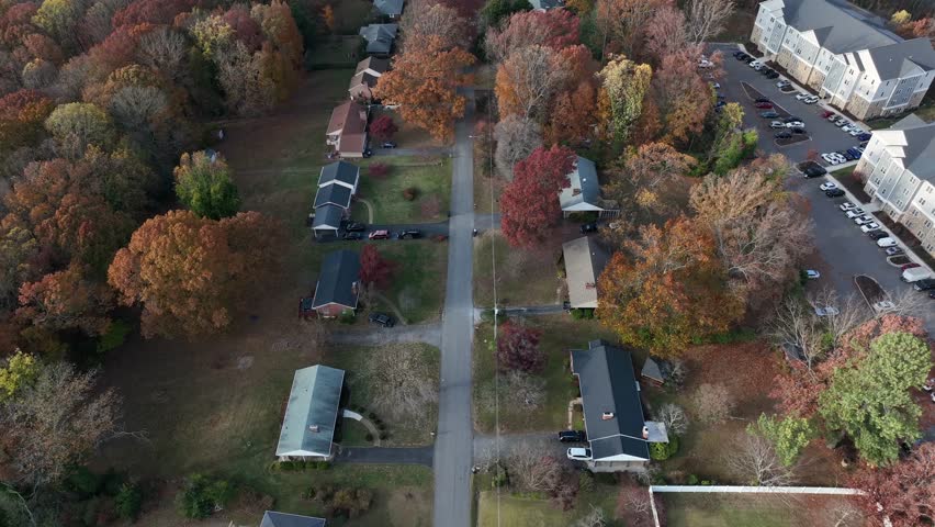 Aerial drone footage showcases an American suburban neighborhood in autumn, with a straight-line flight over a tree-lined street, colorful foliage, and houses under clear midday sunlight.