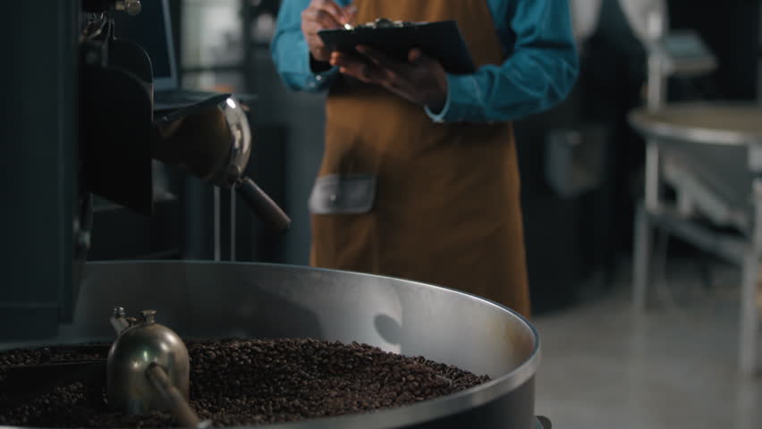Small business owner African American male guy man working in roasted fabric control roasting coffee beans machine process technician checkup machinery equipment checking quality writes notes data - Powered by Shutterstock - Get 15% off with code: PIKWIZARD15