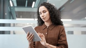 Young serious businesswoman is using digital tablet while standing in a modern business office. Focused beautiful female manager works in application, typing a message or chatting online. Close up - Powered by Shutterstock - Get 15% off with code: PIKWIZARD15