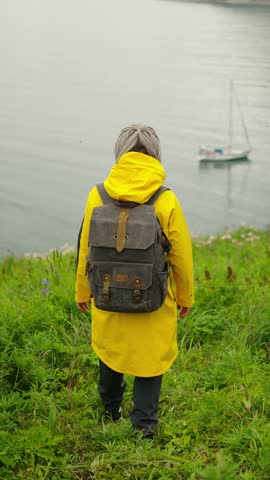 A Journey Through Nature: A Stylish Individual in a Yellow Raincoat and Gray Backpack Gazes at the Tranquil Waters, Embracing the Serene Landscape