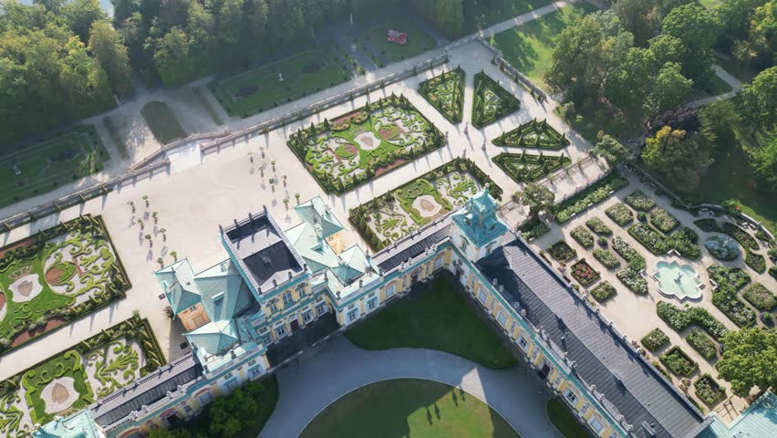 Elegant aerial shot of Warsaw’s Wilanów Palace with decorative sculptures, yellow-white facade, blue roof, and lush park featuring fountain and greenery.